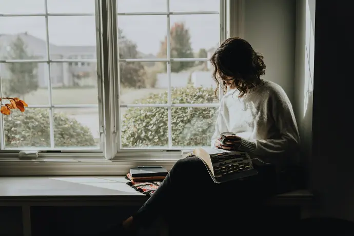A woman sitting on a window sill reading a book